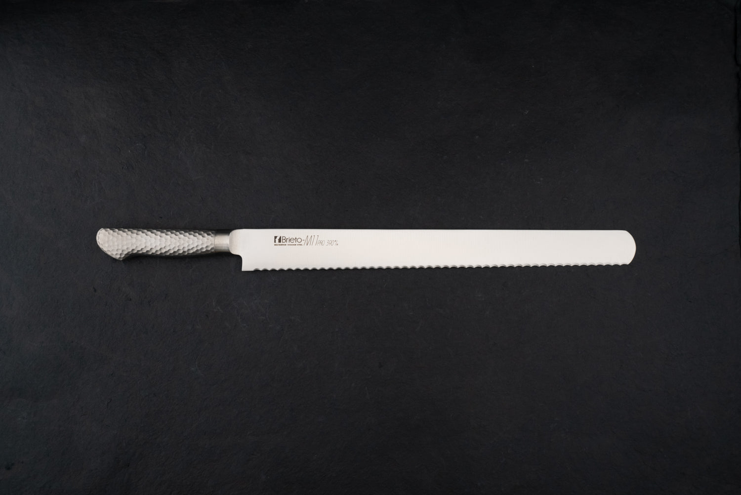 Brieto Pro Bread Knife 390mm M1148 with a long, thin serrated blade and hammered-metal handle, expertly designed for slicing bread without damaging its texture, displayed on a dark background.