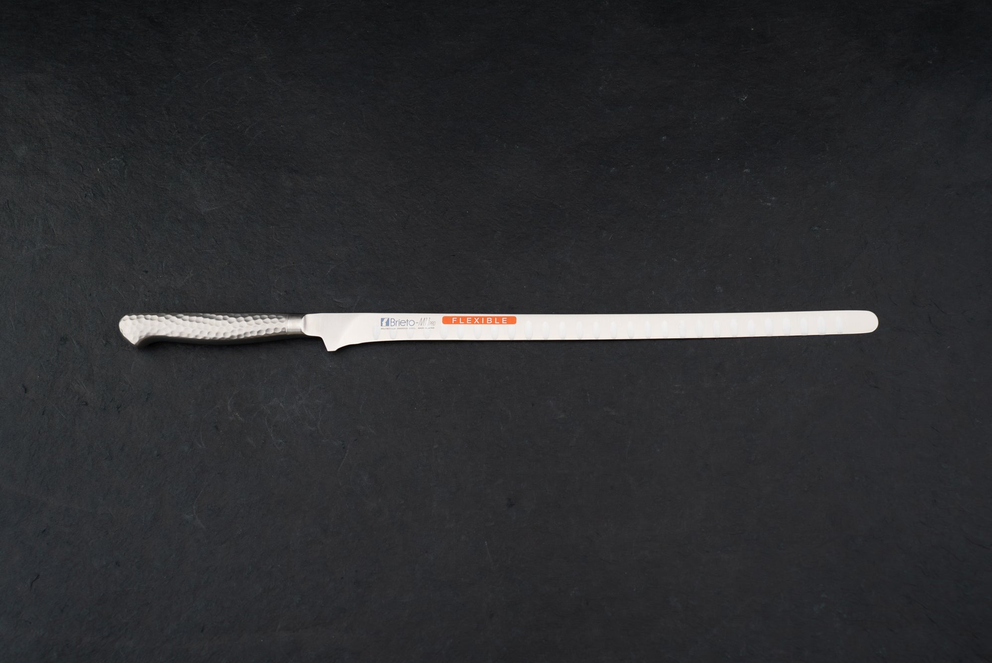 Brieto Pro Flexible Salmon Slicer with Dimples 320mm M116, featuring a long, narrow, flexible blade and hammered-finish metal handle, ideal for precise slicing of meats and fish, displayed on a dark surface.