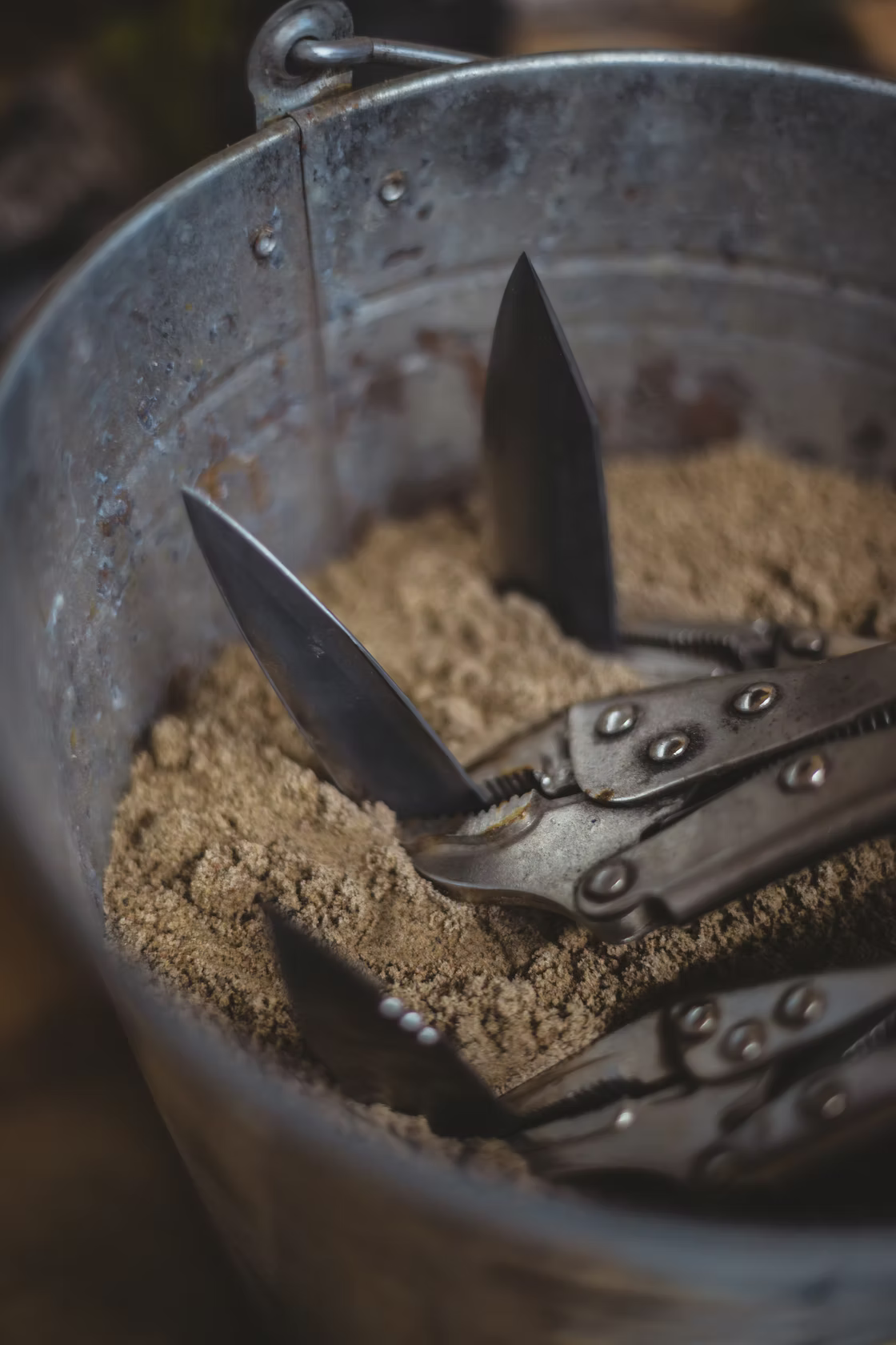 Resqknives Korpi Camp Kitchen Knife with folding blades, crafted from Finnish Birch and recycled stainless steel, displayed upright in a weathered metal pail filled with sand.