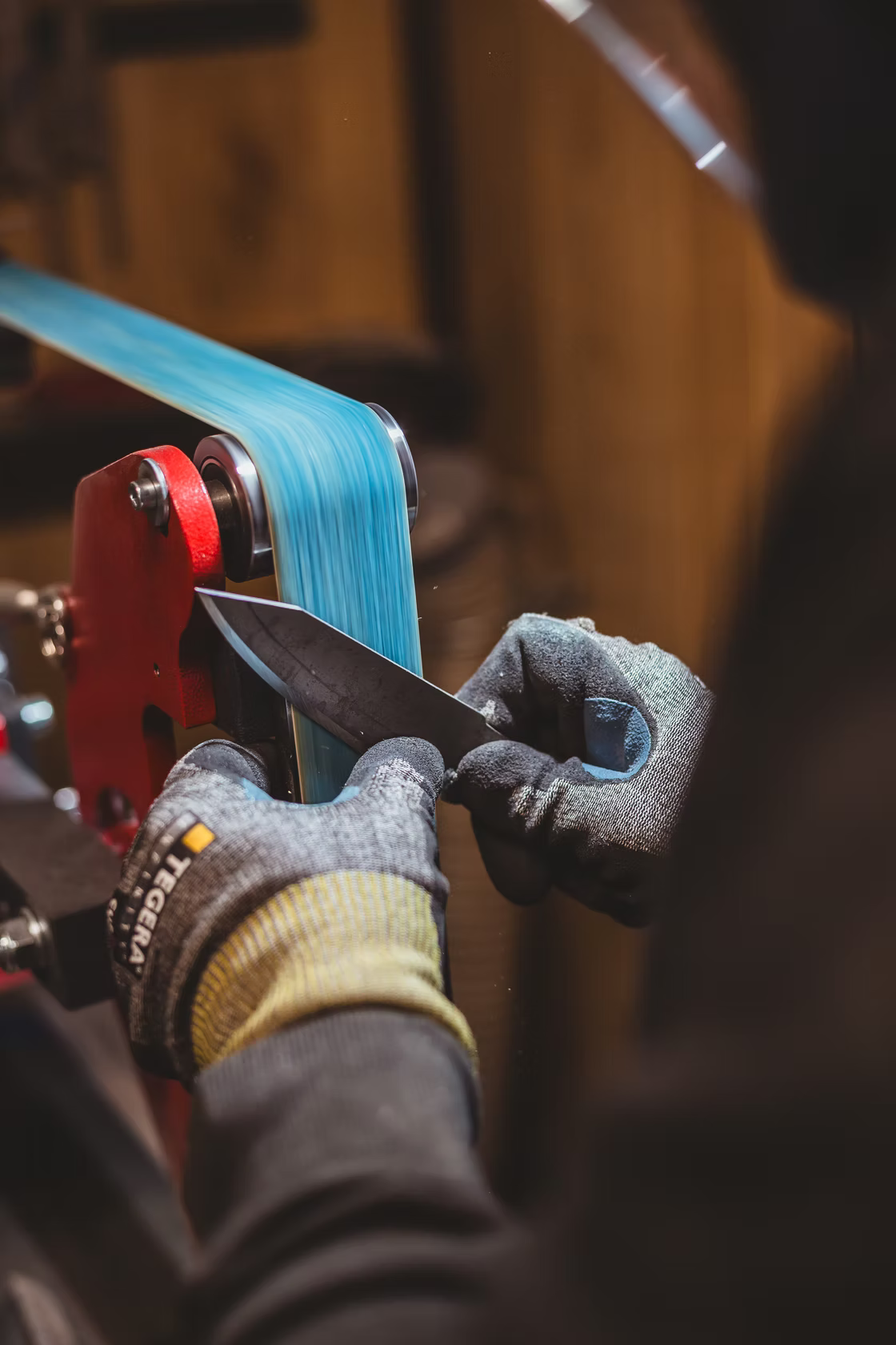 Gloved hand sharpens Resqknives Korpi Camp Kitchen Knife on a belt sander, showcasing the high-quality Finnish Outokumpu stainless steel blade.
