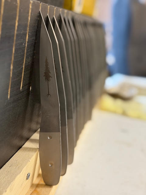 Row of unfinished Resqknives Korpi Camp Kitchen Knife blades with tree emblem, resting on a wooden rack in a workshop, awaiting final crafting stages.